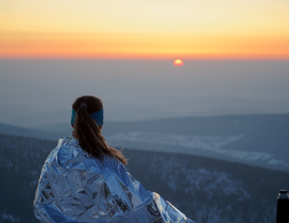 Apifier Míśa wrapped in a termic blanket watching the sun rise outdoors.