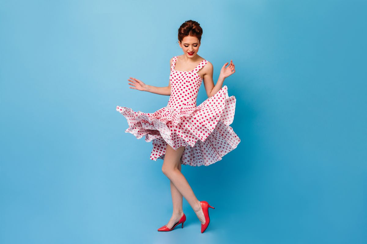 Woman standing in a dancing pose in a white red-dotted dress and red heels, on a blue background.